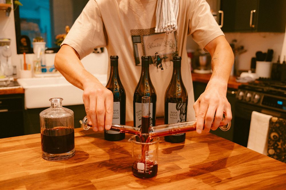 Man using a wine bottle corker on a wooden countertop with multiple wine bottles in a home kitchen.