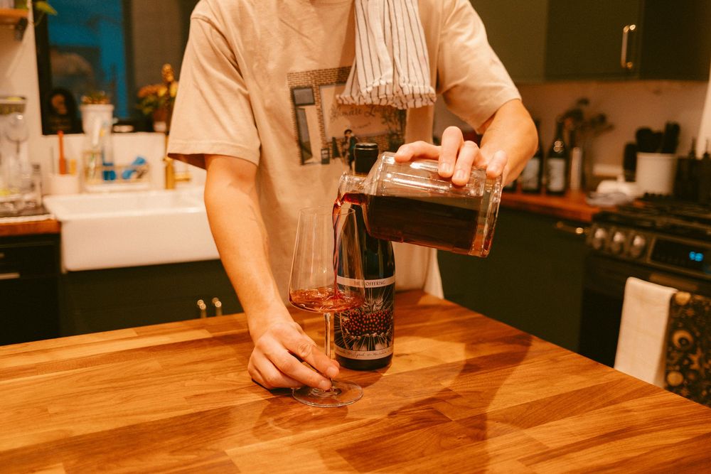 Person pouring red wine from a bottle into a glass decanter on a wooden kitchen counter.