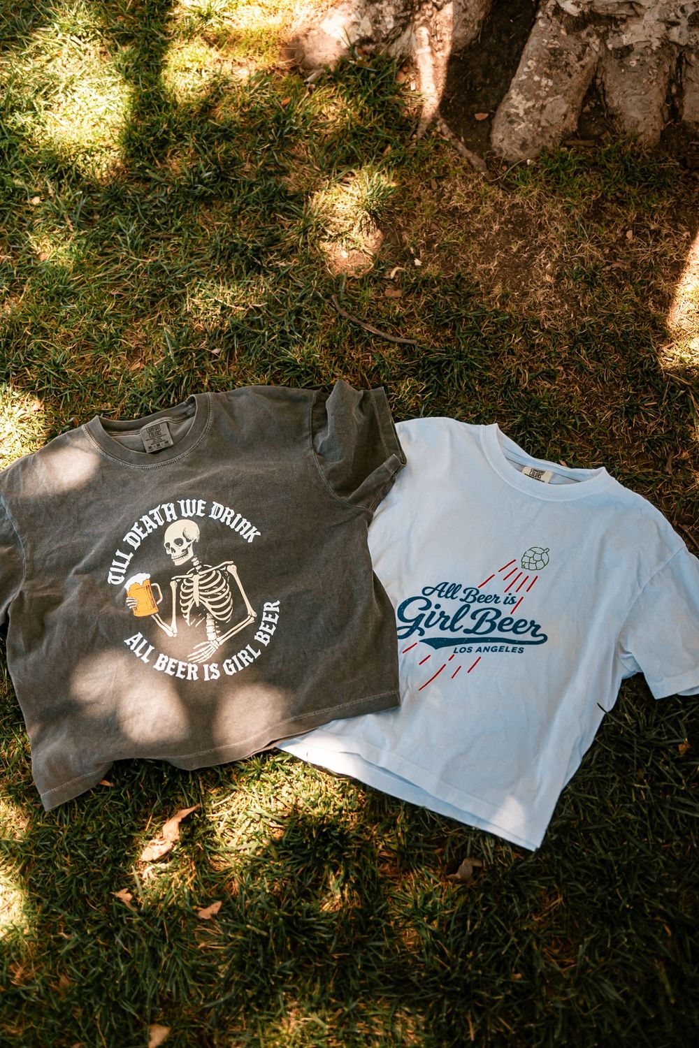 Two graphic tees on grass, one dark with skeleton beer design, one white reading All Beer is Girl Beer.
