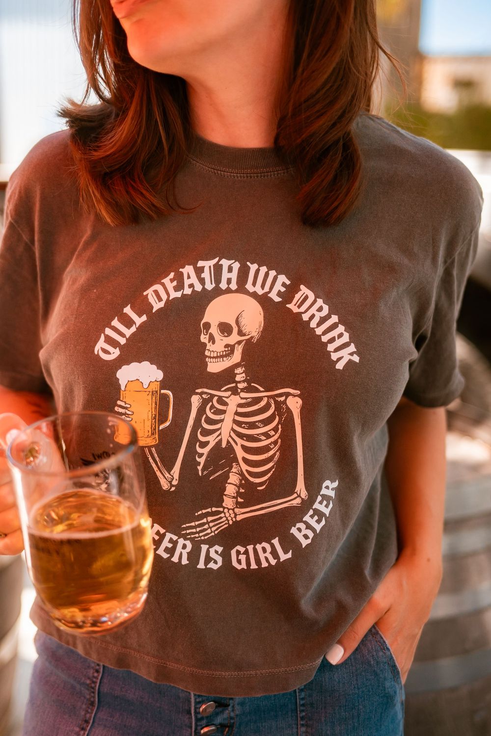 Woman wearing a dark gray skeleton "Till Death We Drink, Beer Is Girl Beer" graphic tee holding a glass of beer.