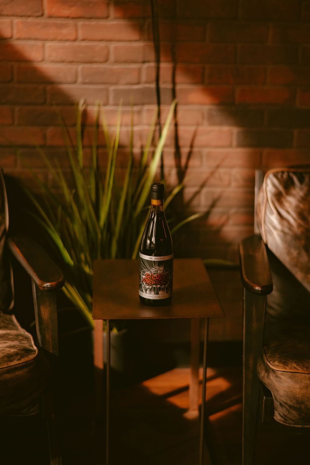 A wine bottle sits on a small wooden table between leather chairs with a brick wall and green plant backdrop.
