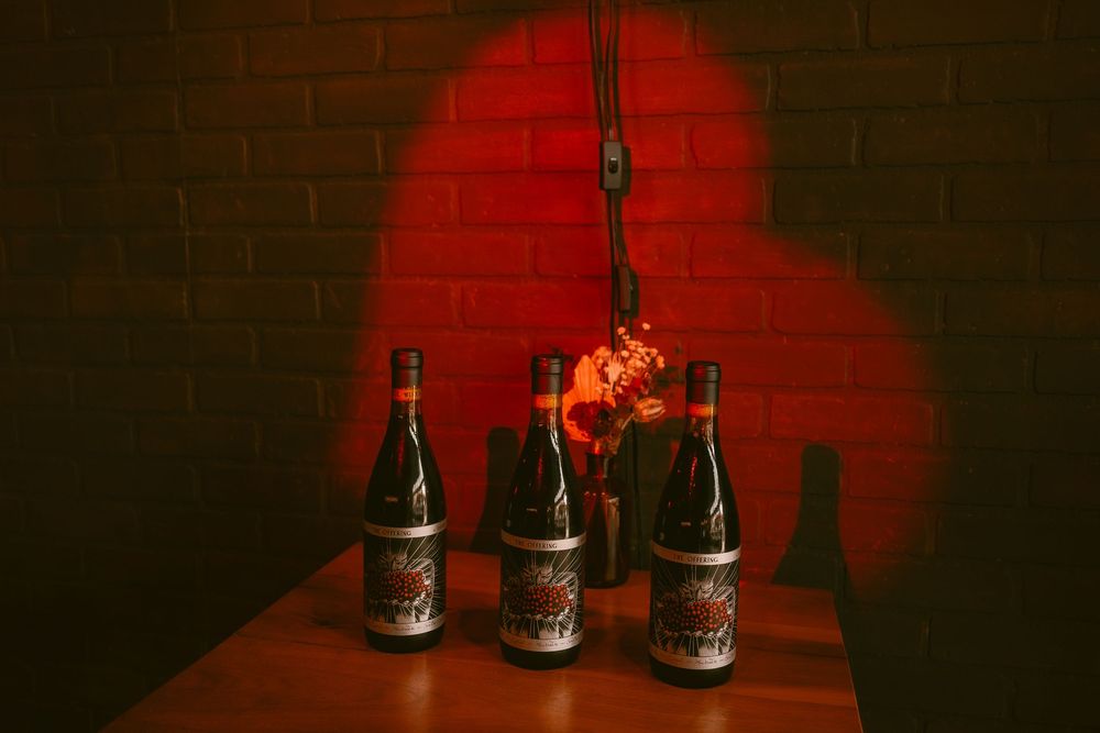 Three wine bottles with decorative labels arranged on a wooden table under dramatic red lighting against a brick wall.