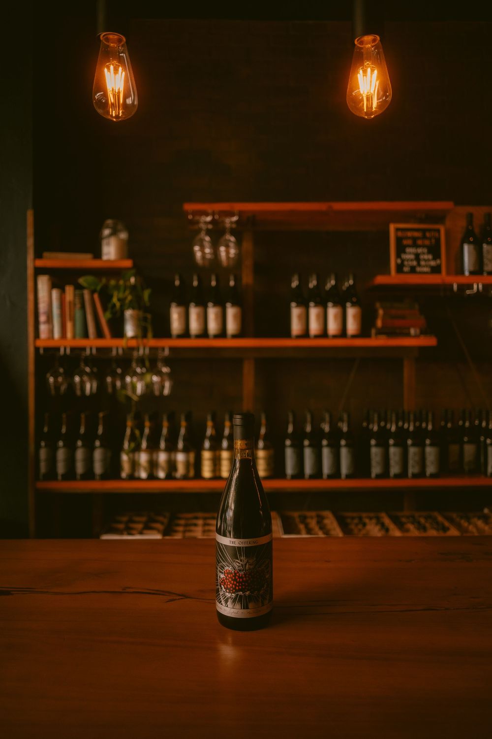 A wine bottle on a bar counter with Edison bulb lighting and shelves of wine bottles in the background.