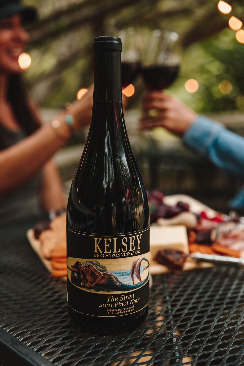 Kelsey Bee Canyon Vineyards The Siren 2021 Pinot Noir wine bottle on outdoor table with charcuterie board and string lights.