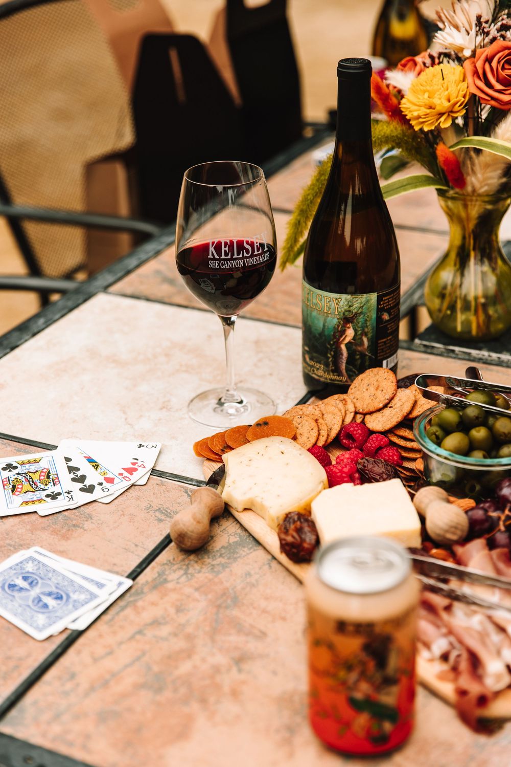 Kelsey Bee Canyon Vineyards wine glass and bottle on tile table with charcuterie board, playing cards, and floral arrangement.