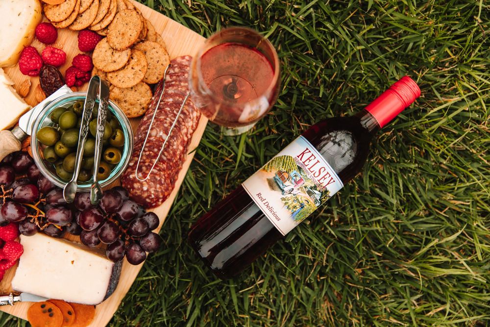 Kelsey wine bottle and rose glass on grass beside a wooden charcuterie board with cheese, grapes, and crackers.