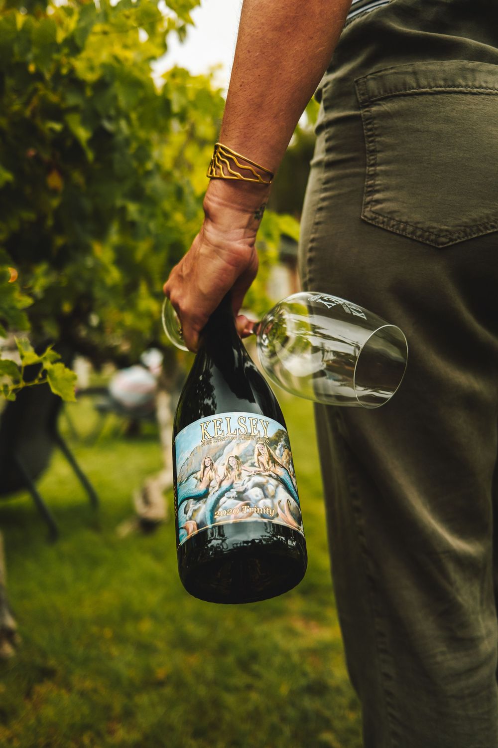 Person holding Kelsey wine bottle and empty glass while standing in a lush green vineyard.