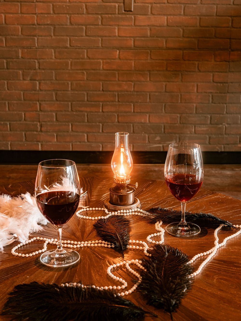 Two glasses of red wine on a wooden table with pearl necklaces, feathers, and a glowing oil lamp against a brick wall.