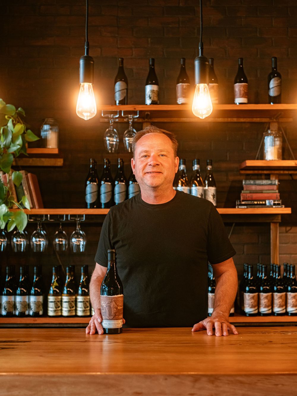 Man in black shirt standing behind a wooden bar with wine bottles and Edison bulb lighting in a cozy wine shop.