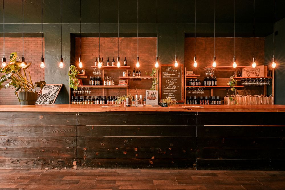 Rustic bar interior with Edison bulb lighting, dark wood counter, and shelves lined with bottles.