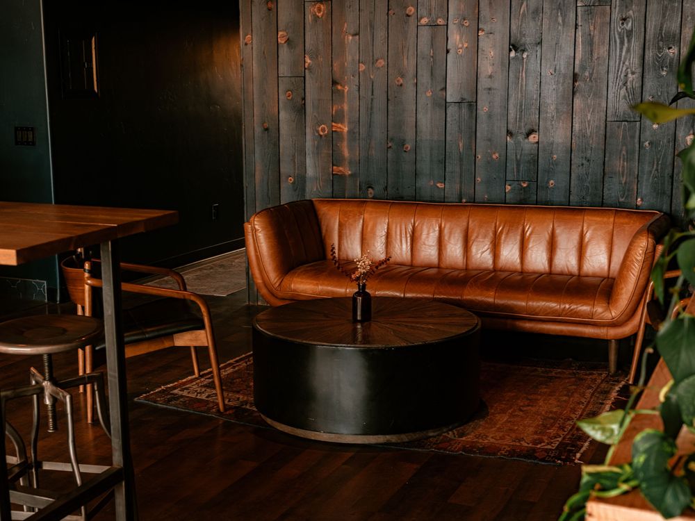 Brown leather curved sofa with black round coffee table on dark wood floor against rustic teal wood panel wall.