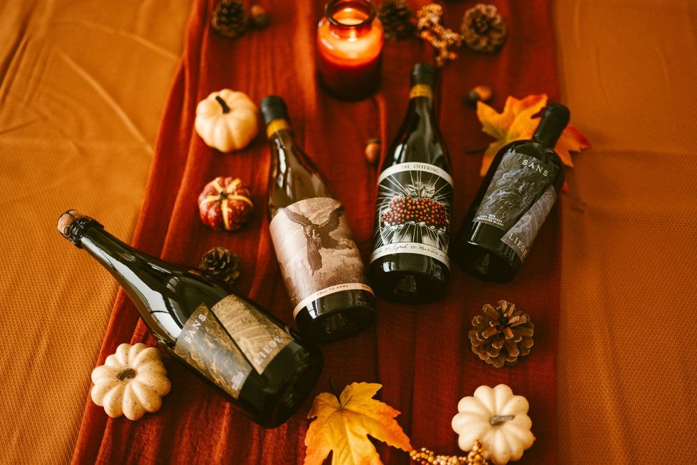 Four Kelsey wine bottles displayed on rustic red fabric with mini pumpkins, pine cones, and autumn leaves.