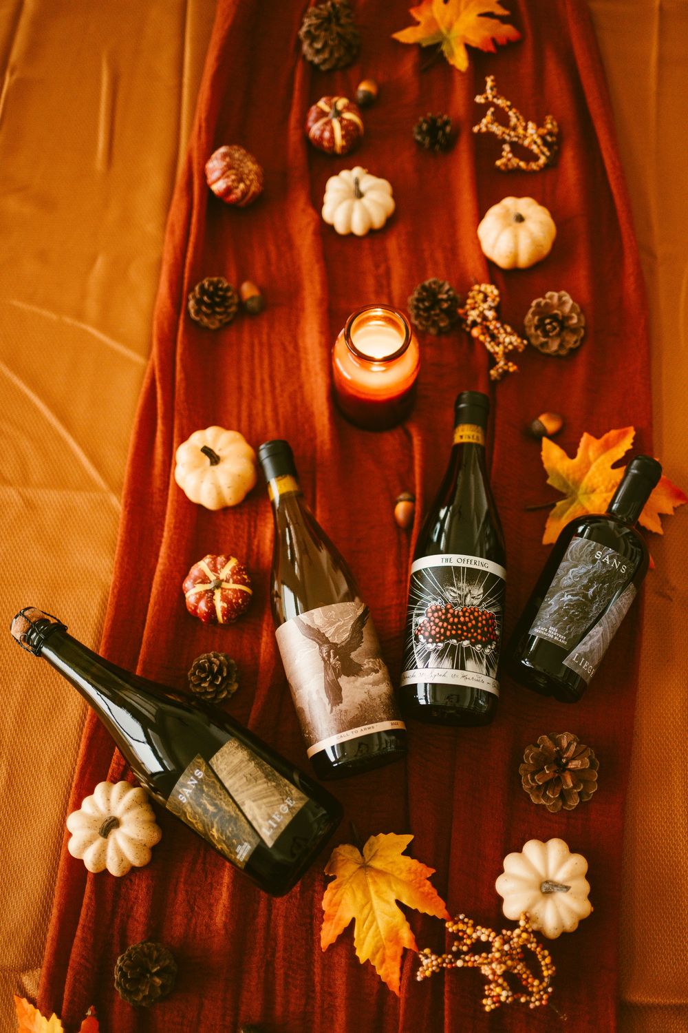 Four Kelsey wine bottles arranged on red fabric surrounded by white pumpkins, fall leaves, and a glowing candle.