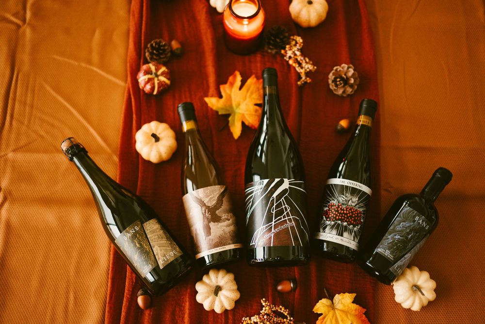 Four Kelsey wine bottles laid on red draped fabric with autumn decorations including mini pumpkins and maple leaves.