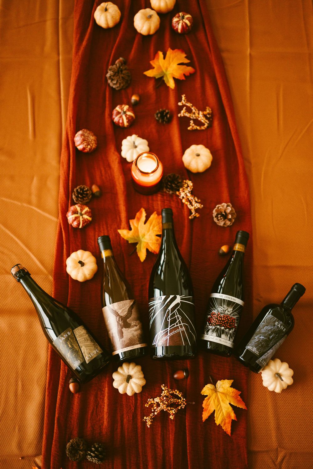 Five Kelsey wine bottles displayed on red fabric with fall seasonal decor including pumpkins, leaves, and a candle.