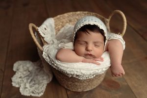 Newborn baby sleeping peacefully in a wicker basket with white lace blanket and knit bonnet on wood floor.
