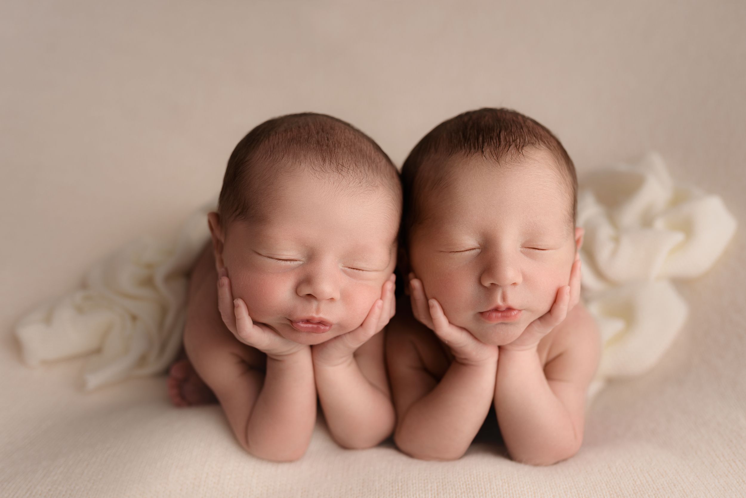 Two sleeping newborn twins posed side by side with hands under chins on a soft beige blanket with white flowers.