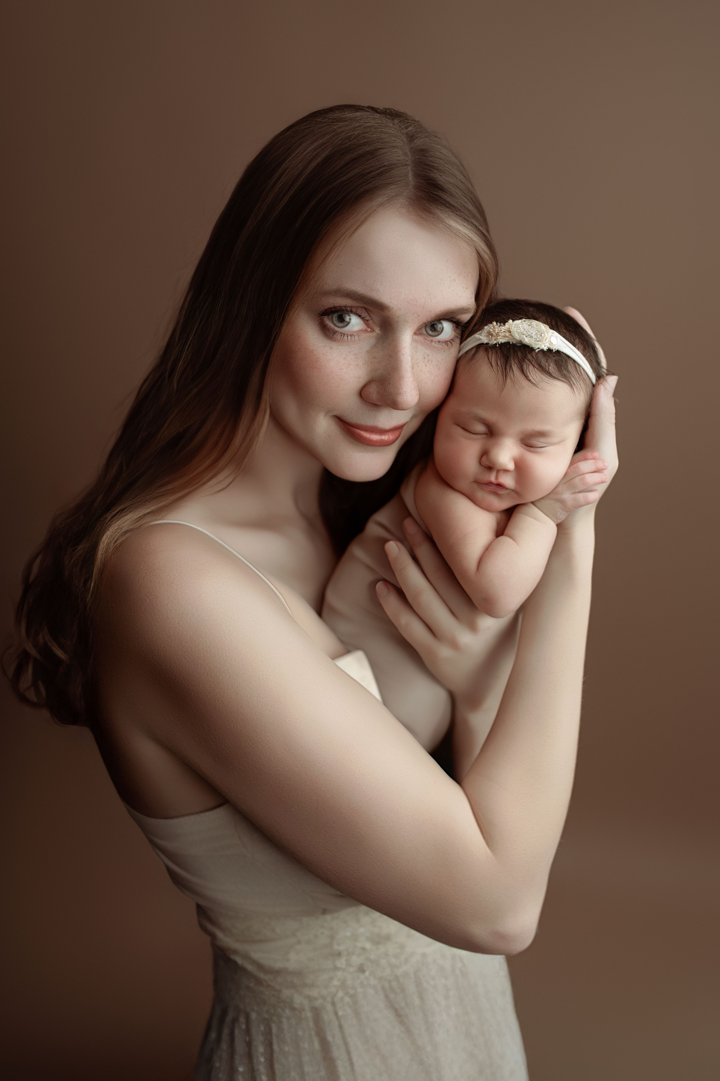 A tender portrait of a mother in a cream dress holding her sleeping newborn baby against a brown background.