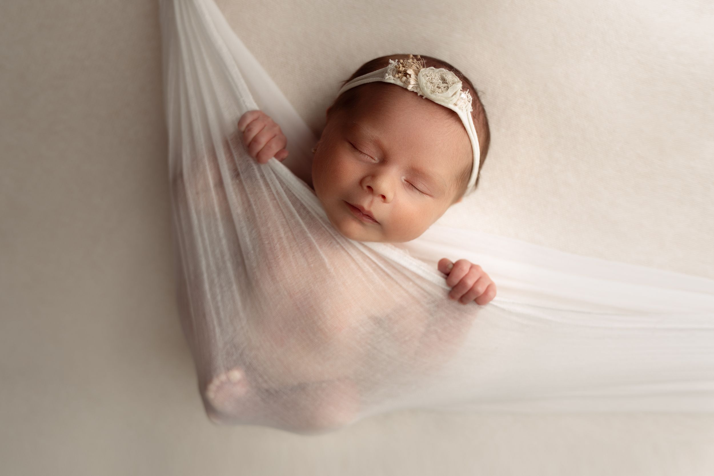 Sleeping newborn cradled in delicate white fabric hammock with lace headband.