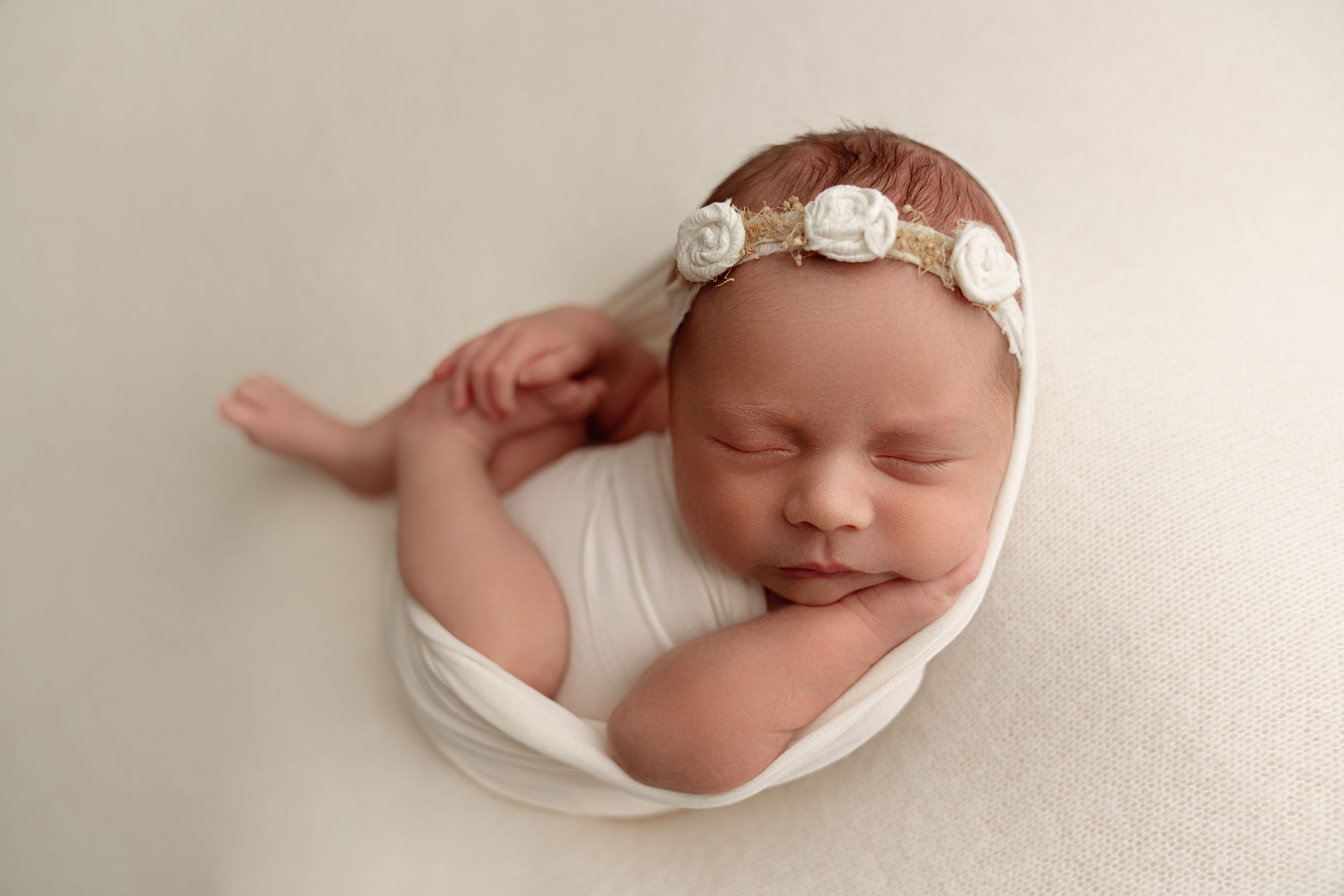 Serene newborn wearing floral headband and white wrap resting on neutral backdrop.