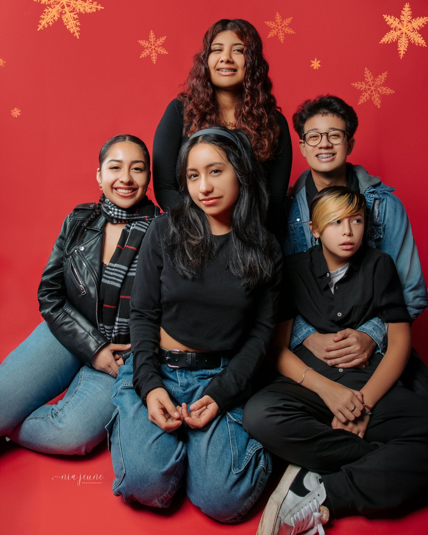 People in casual denim and black outfits pose together against a festive red background with gold snowflakes.