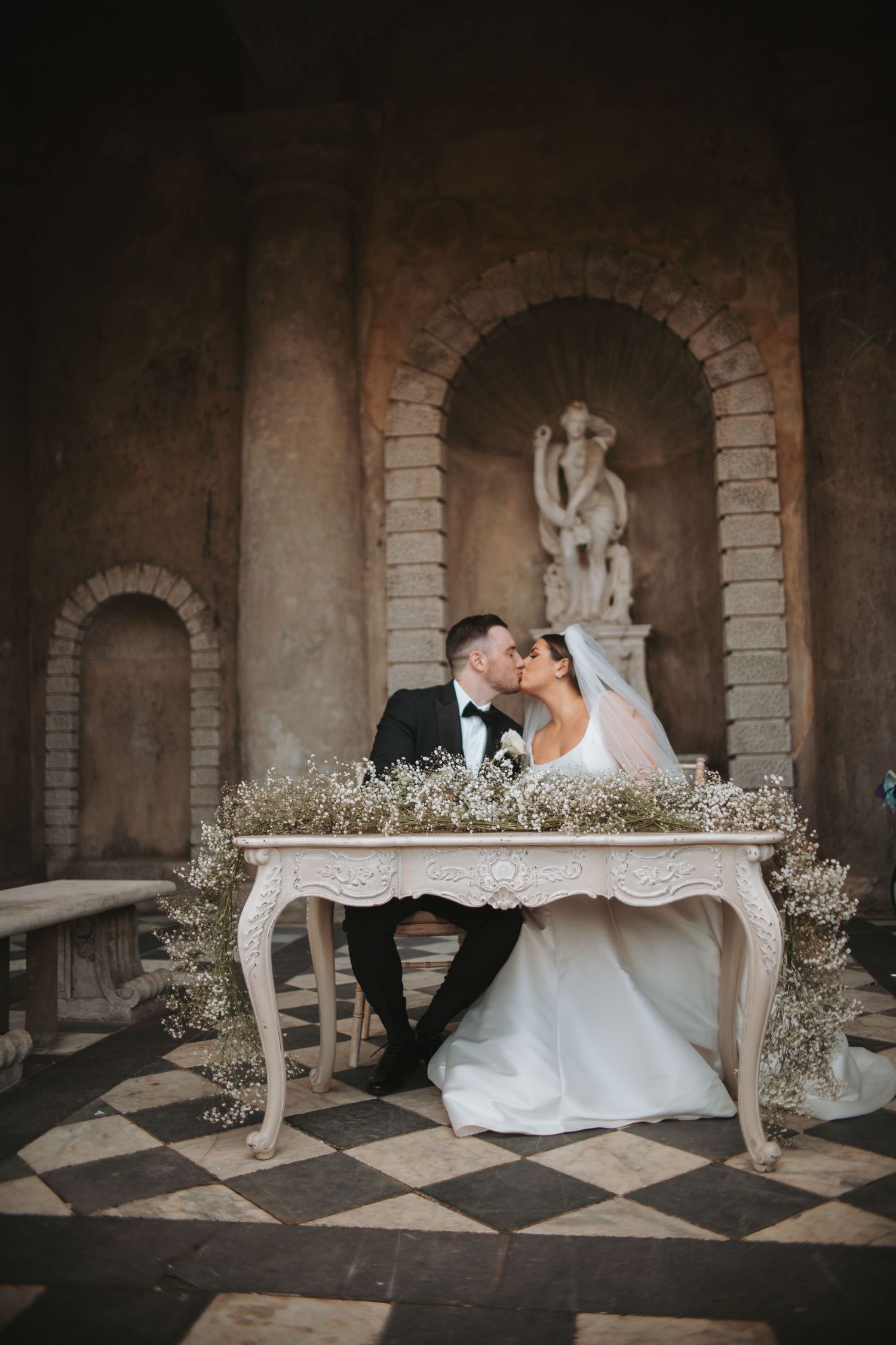 Couple shares intimate moment at ornate vintage table with floral decor and classical statue in background.