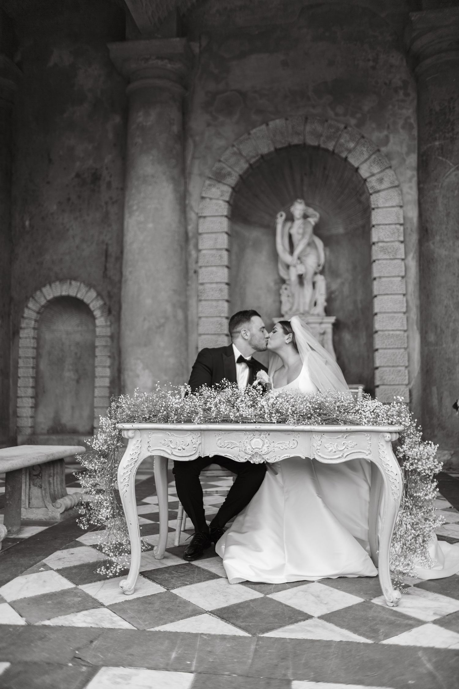 Black and white portrait of newlyweds at elegant table with romantic archway setting.