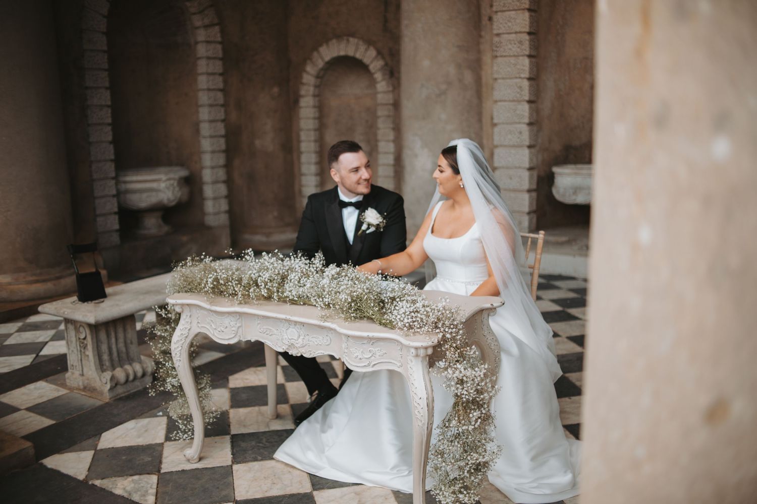 Wedding couple poses at decorative table adorned with baby's breath in classical architectural setting.