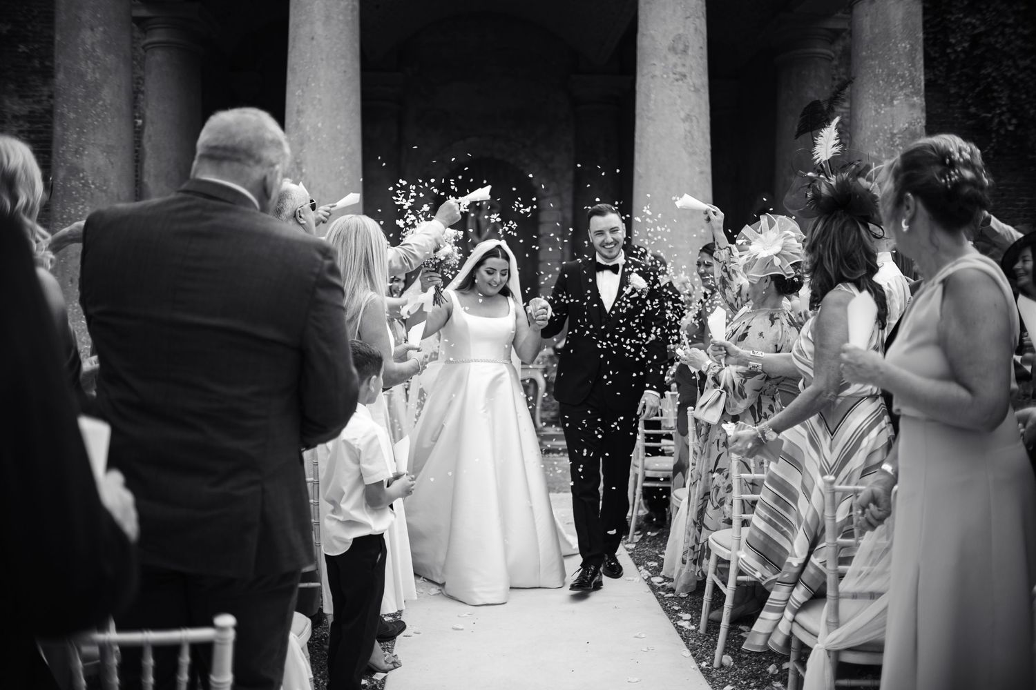 Wedding guests throw confetti as couple walks down outdoor colonnade aisle.