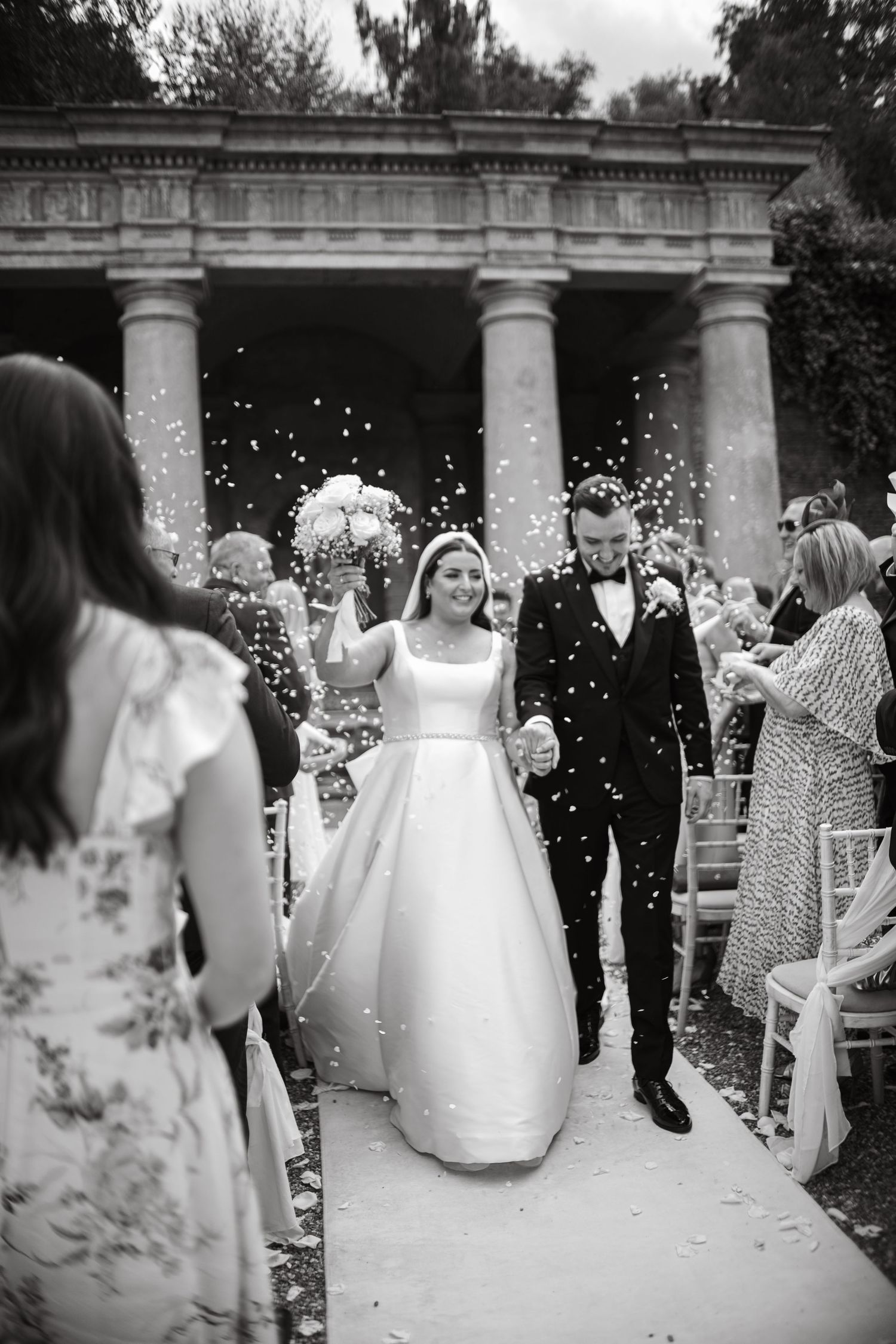 Elegant couple walks down aisle while guests shower them with confetti under columned structure.