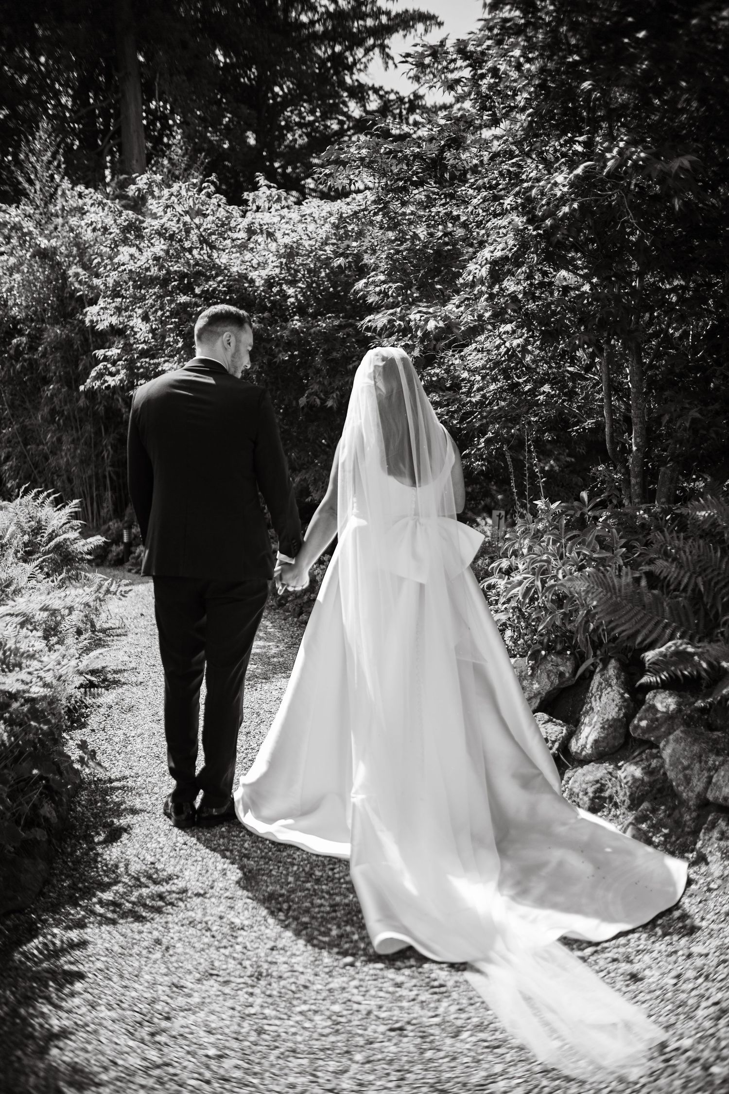 Black and white photo of couple walking down garden path with flowing wedding dress train.