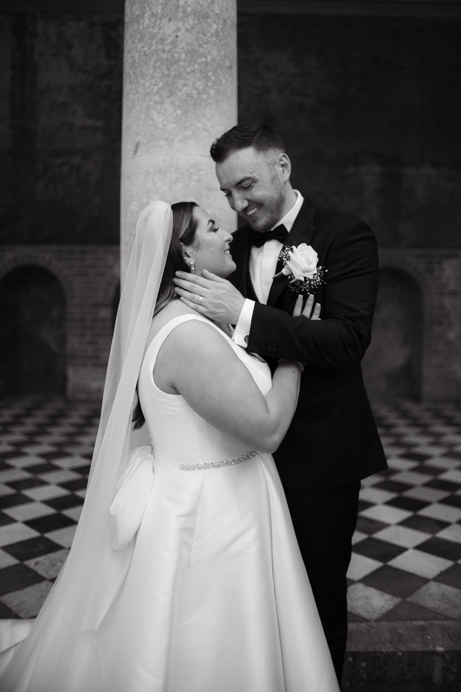 Intimate black and white portrait of couple embracing against checkered floor and stone columns.