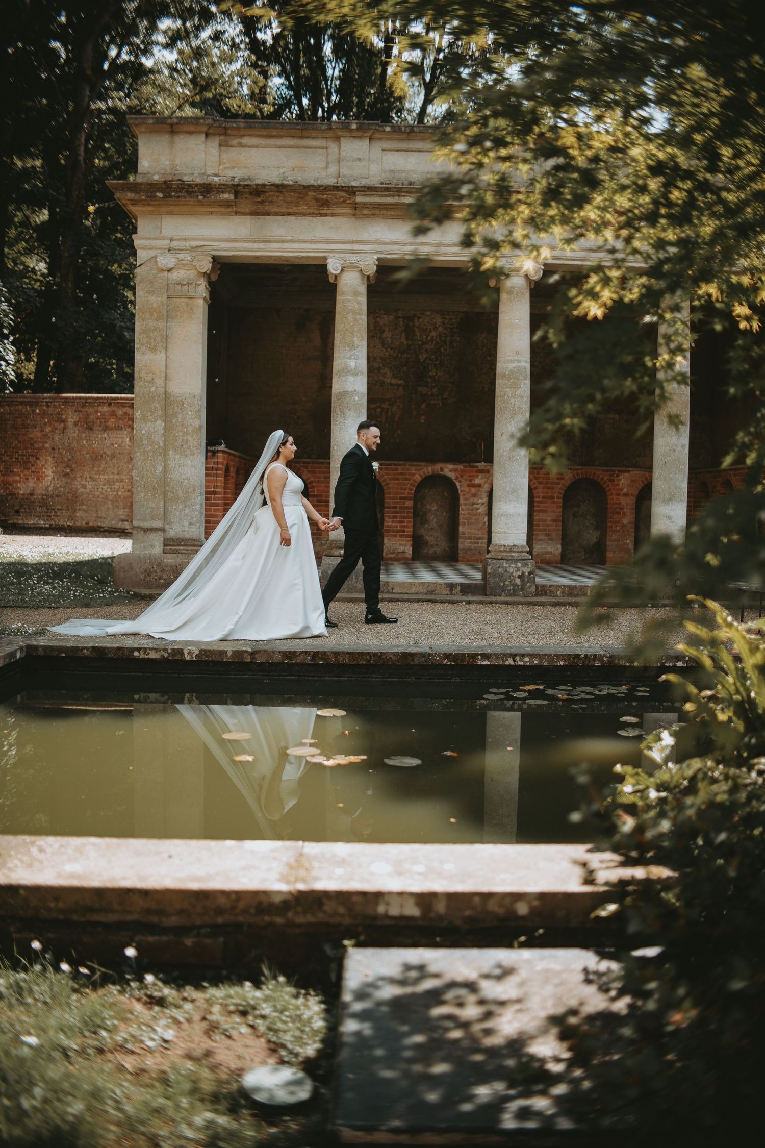 Wedding couple walks beside ornate stone colonnade and reflecting pool in garden setting.