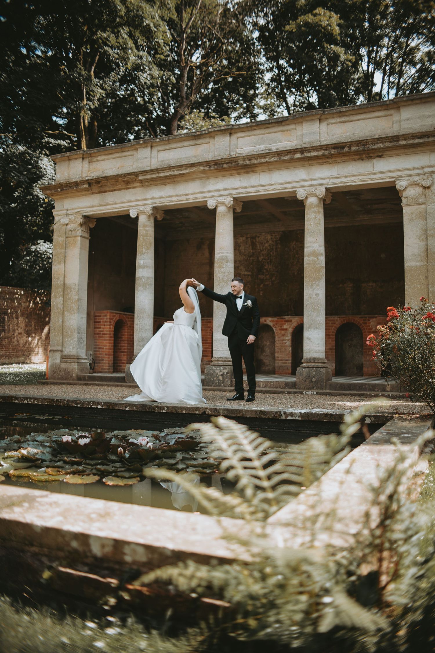 Couple shares intimate moment under grand stone colonnade with flowering bushes in foreground.