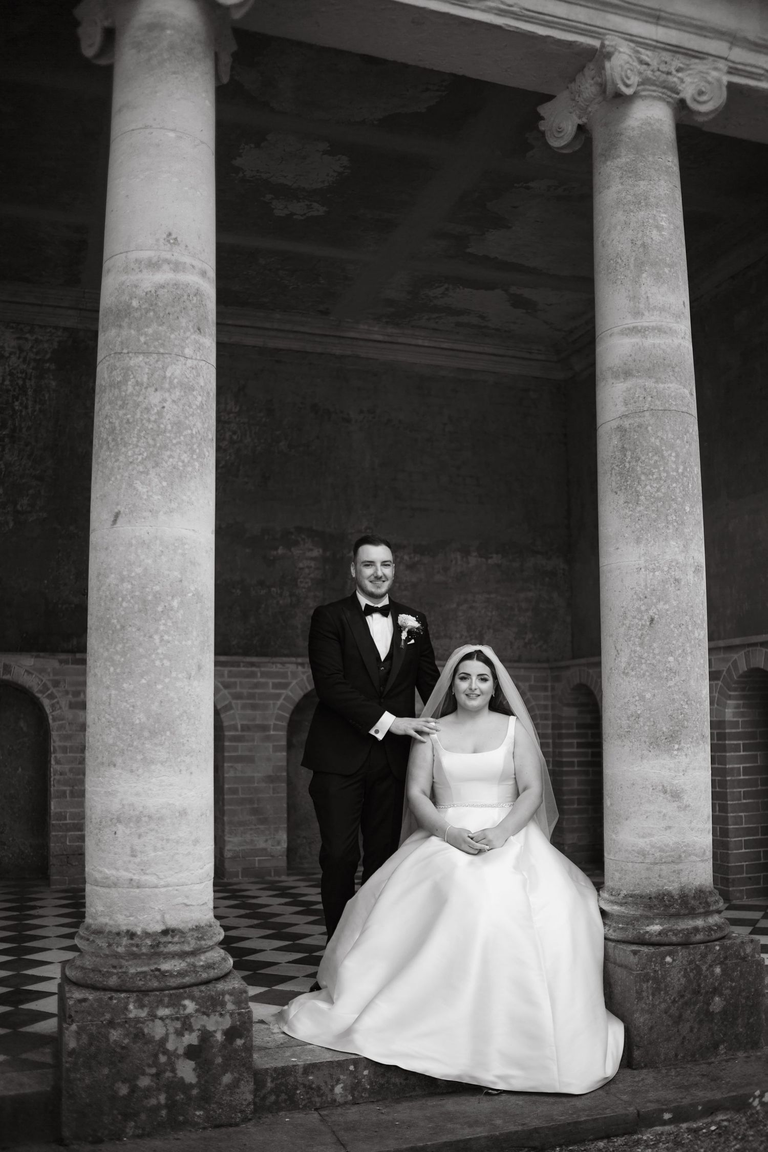 A wedding couple poses elegantly between classical stone columns in dramatic black and white photography.