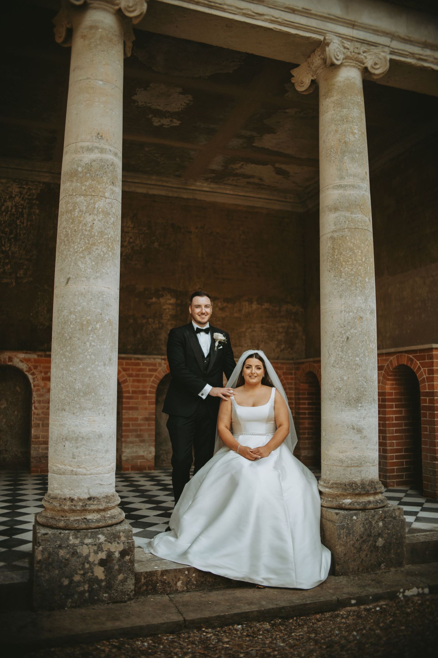 Newlyweds pose between ornate stone columns in a historic architectural setting.