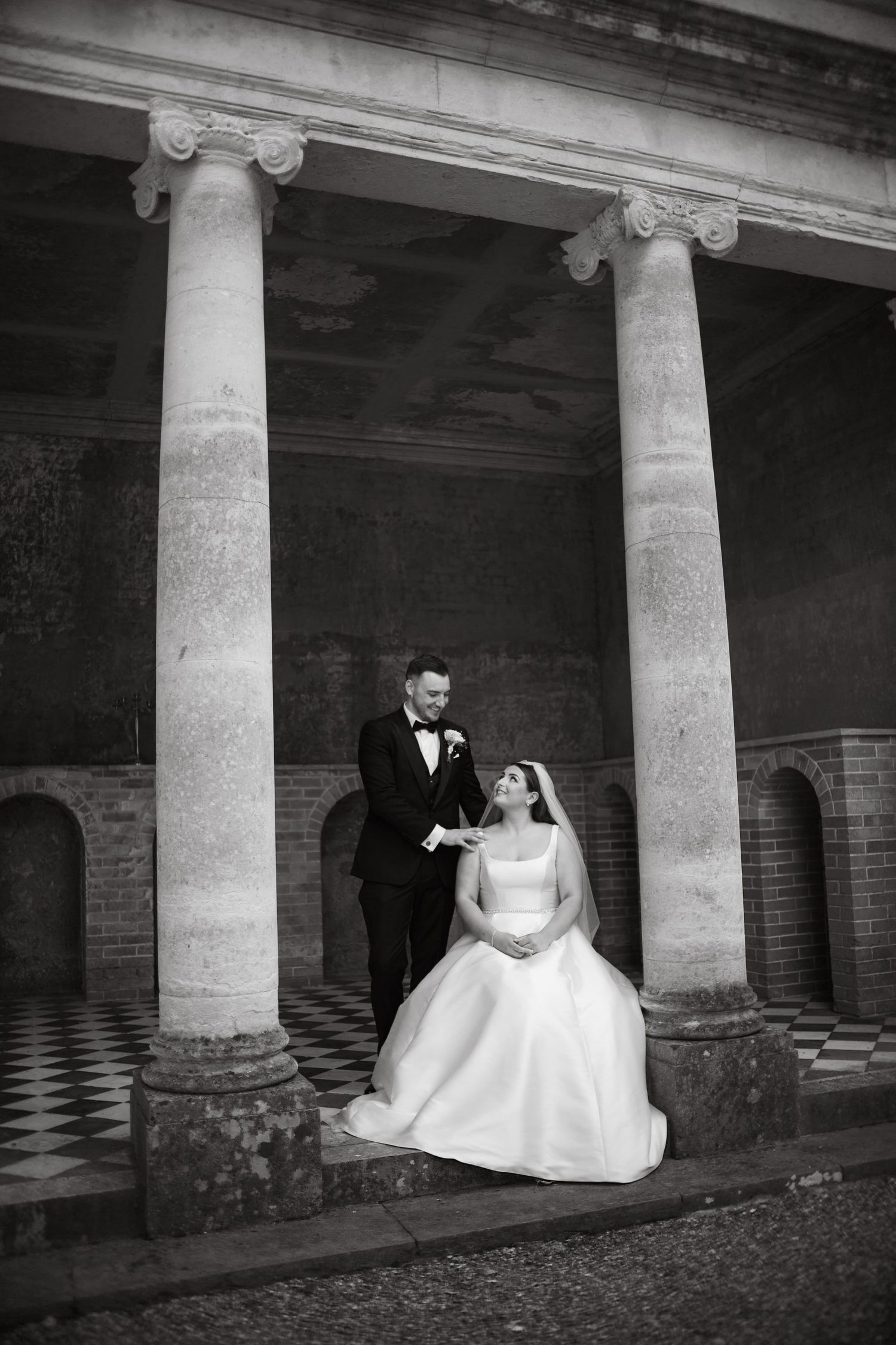 Artistic black and white wedding portrait featuring a couple among classical stone pillars.