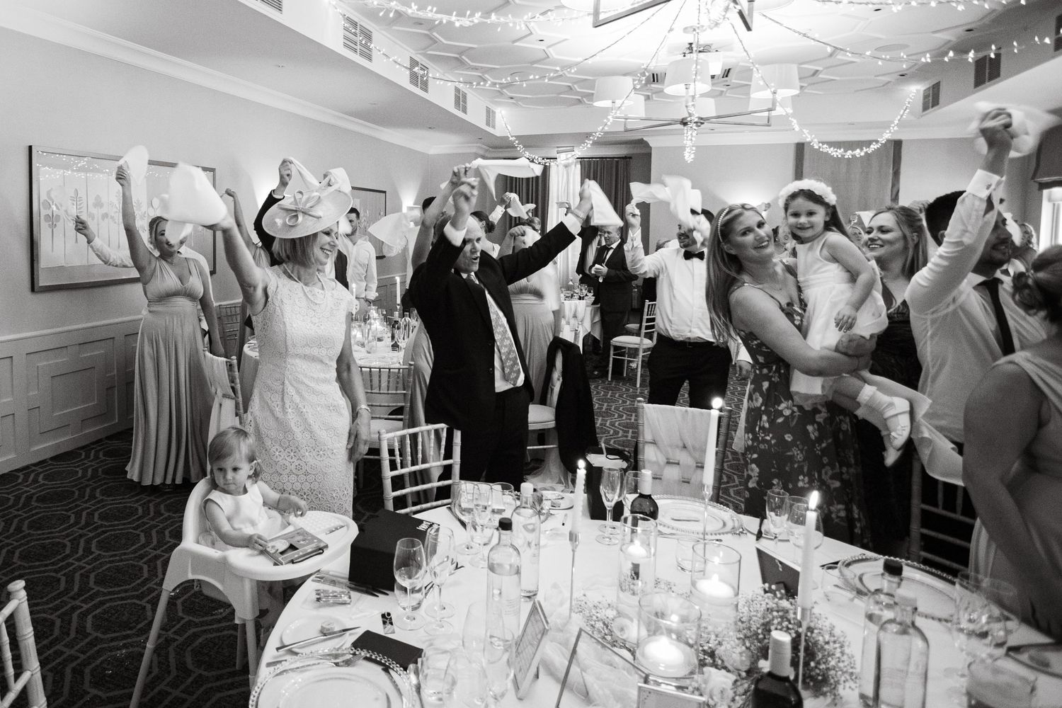 Black and white photo of wedding guests celebrating with raised arms in a reception hall.