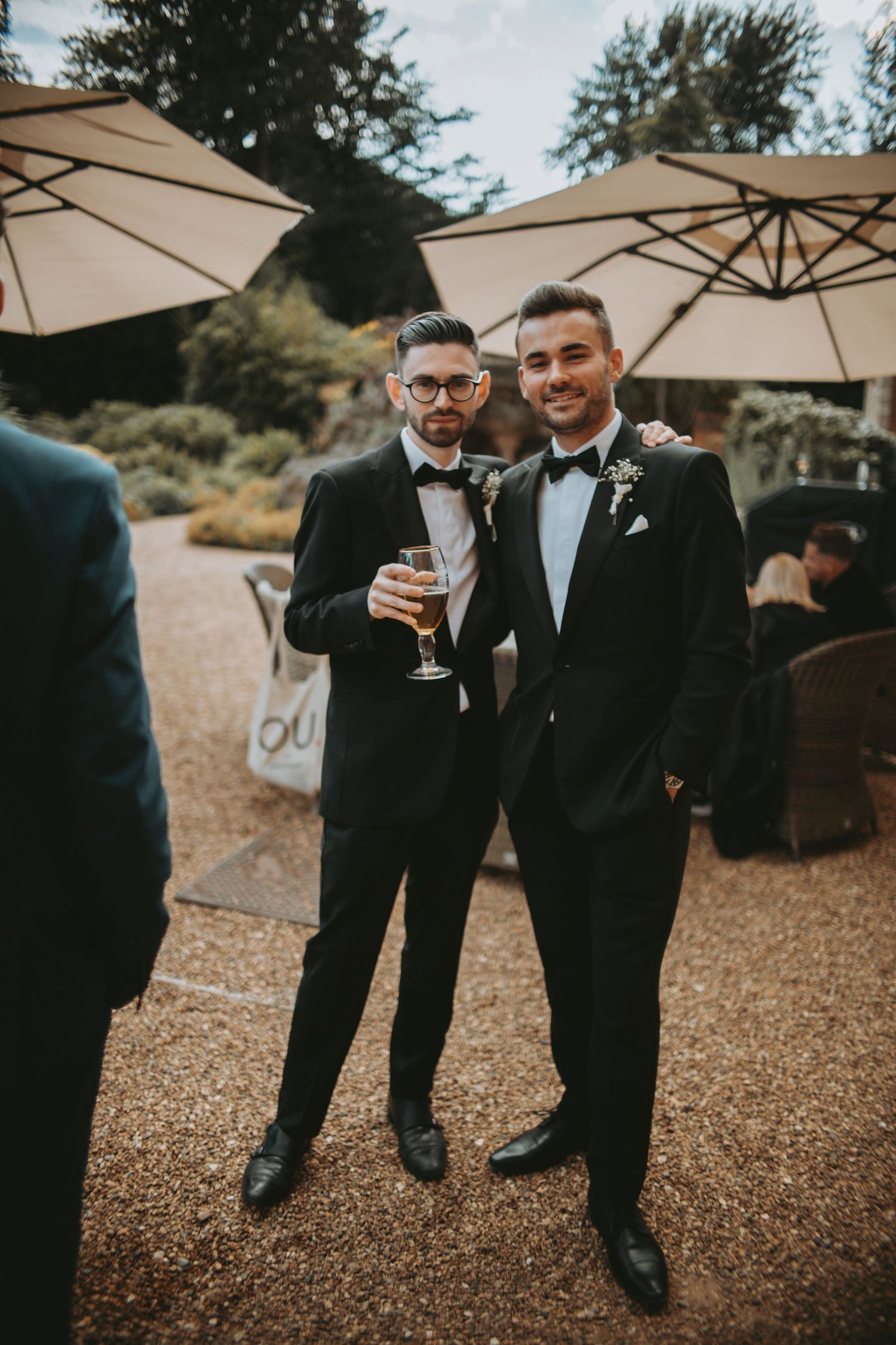 Groomsmen in formal black tuxedos share drinks and conversation under cream-colored patio umbrellas.