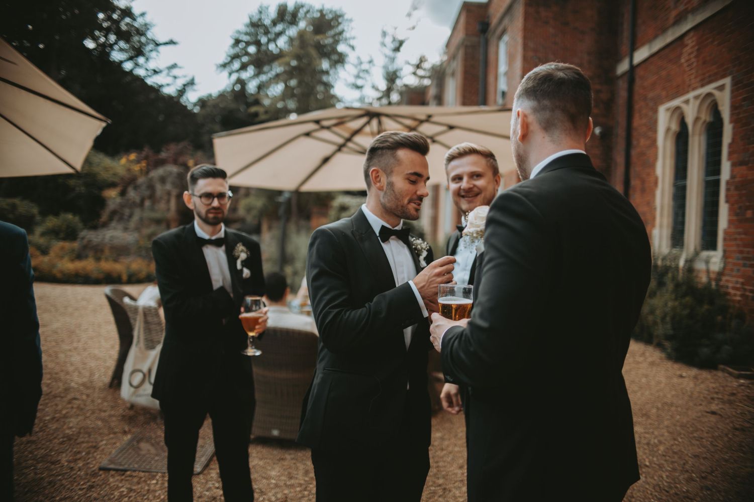 Wedding party members in black tuxedos gather outdoors under beige umbrellas for a celebration.
