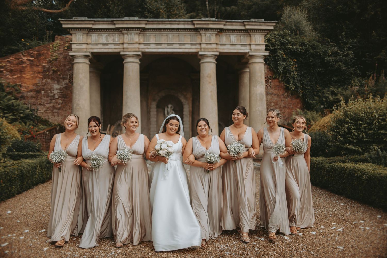Bridal party in neutral dresses poses with bride in white gown under ornate stone colonnade.