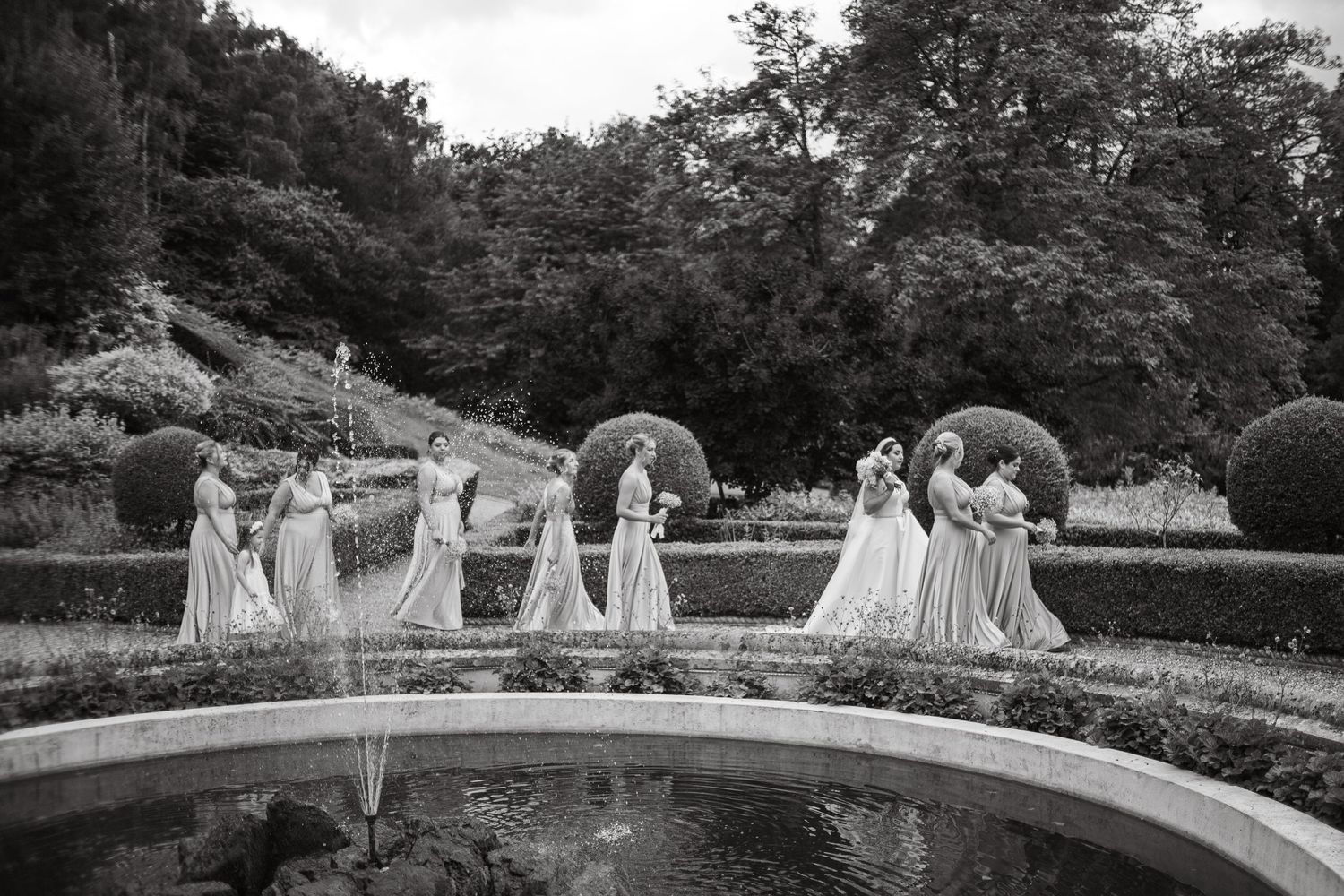 Wedding party members walk along curved stone wall in garden setting.