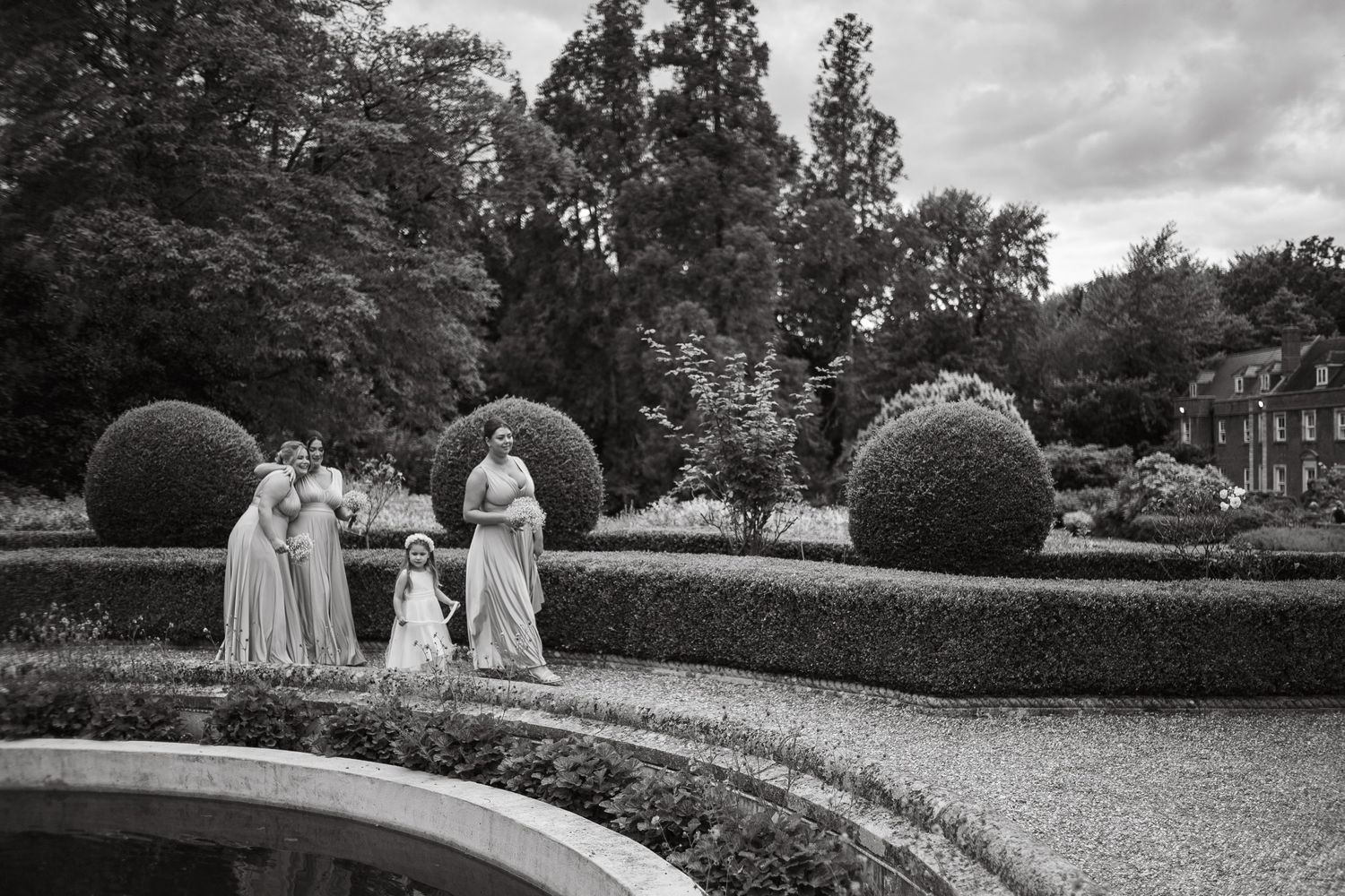 A black and white photo of figures in formal attire walking through an elegant garden with manicured hedges.