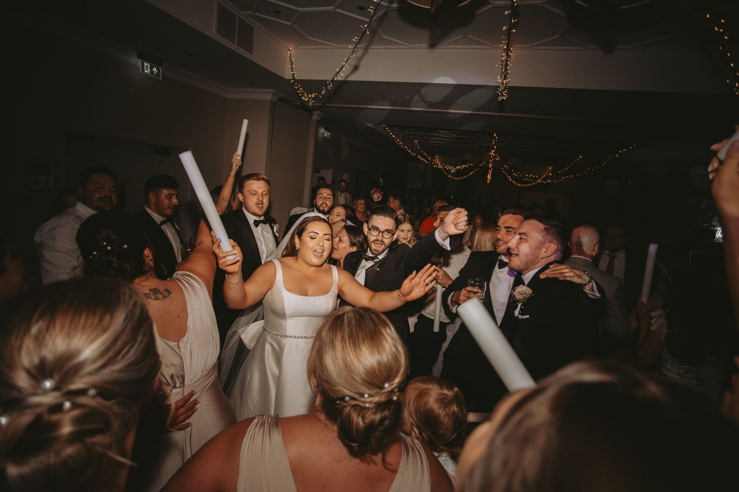 Energetic wedding reception party with crowd dancing under dim lighting and decorative elements.