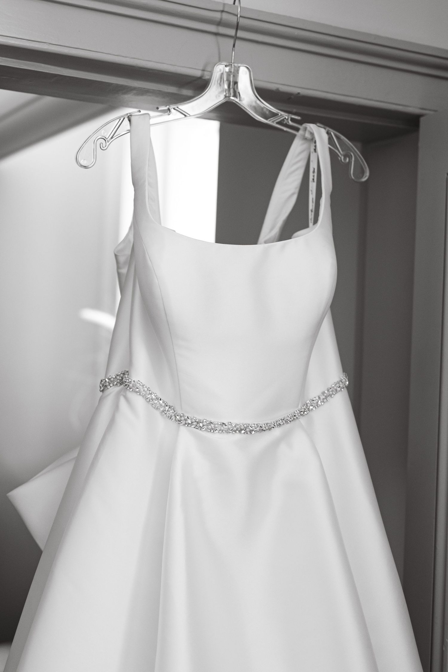 Elegant white wedding dress with crystal detail hanging against neutral wall.