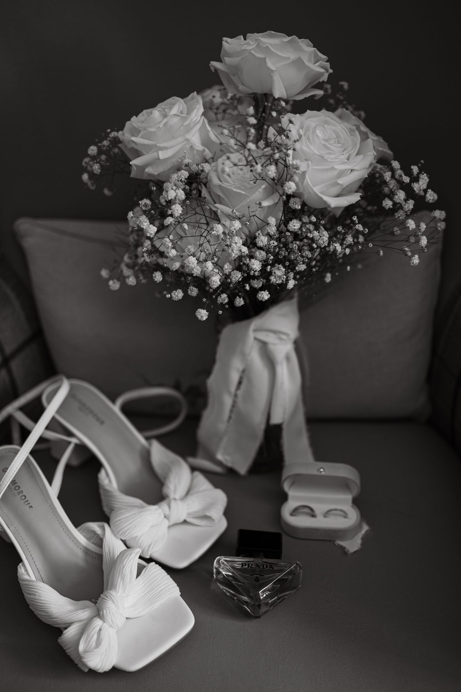 Wedding details including white roses, bridal shoes, and rings arranged on a dark surface.