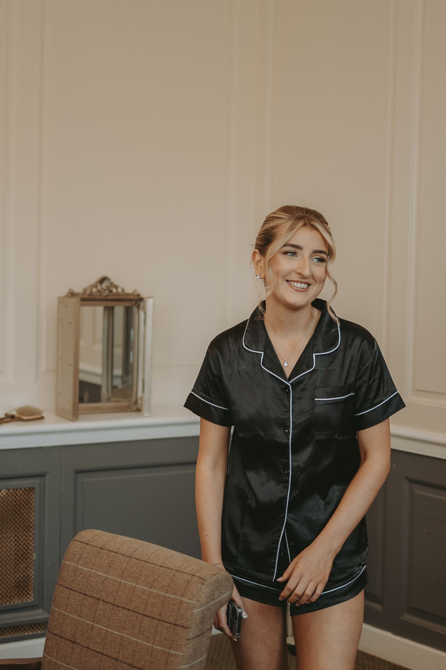 Person wearing black silk pajamas sitting and smiling in an elegant bedroom setting.