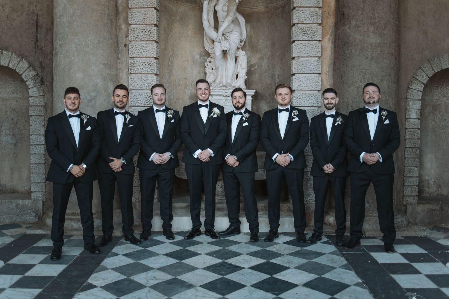 Large wedding party of groomsmen in black tuxedos posed formally against stone architecture.