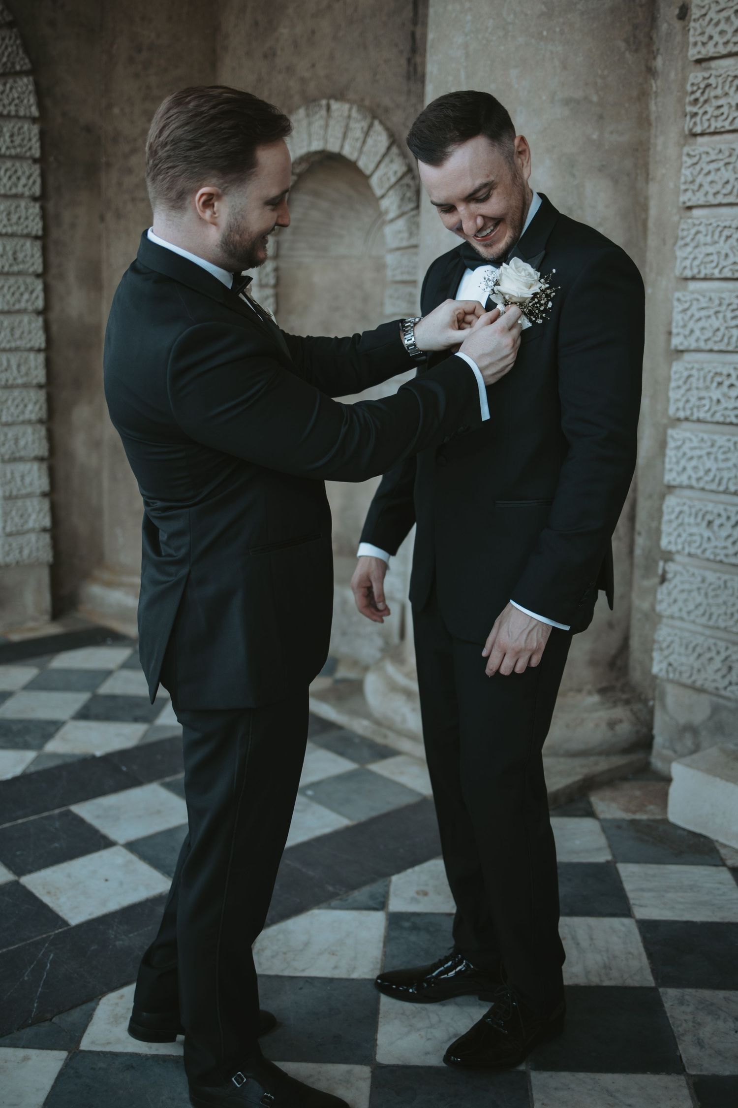 Groomsmen in black tuxedos sharing lighthearted moments in a stone corridor with checkered floor.