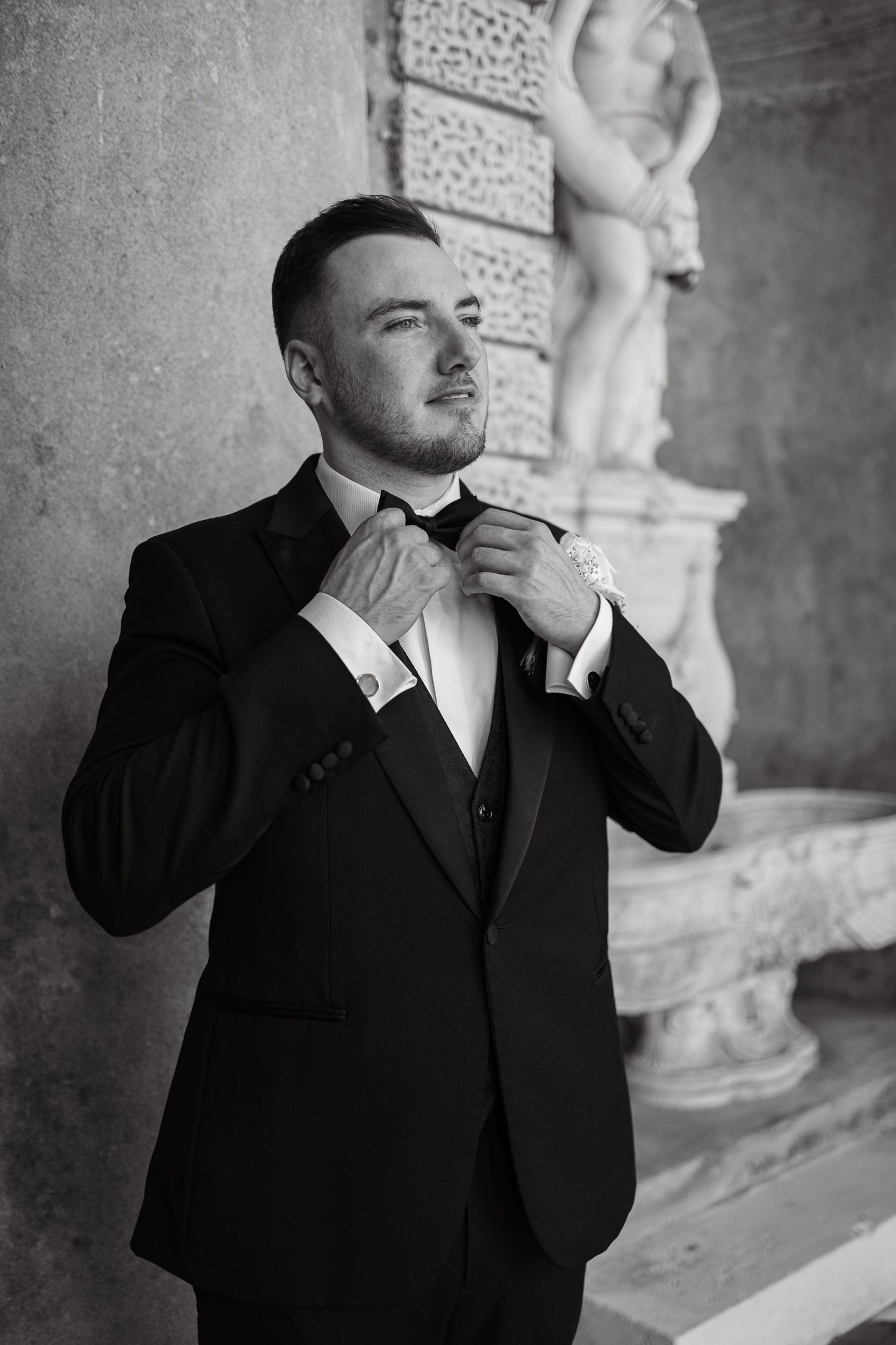 A groom in formal attire adjusts his bowtie near a classical statue.